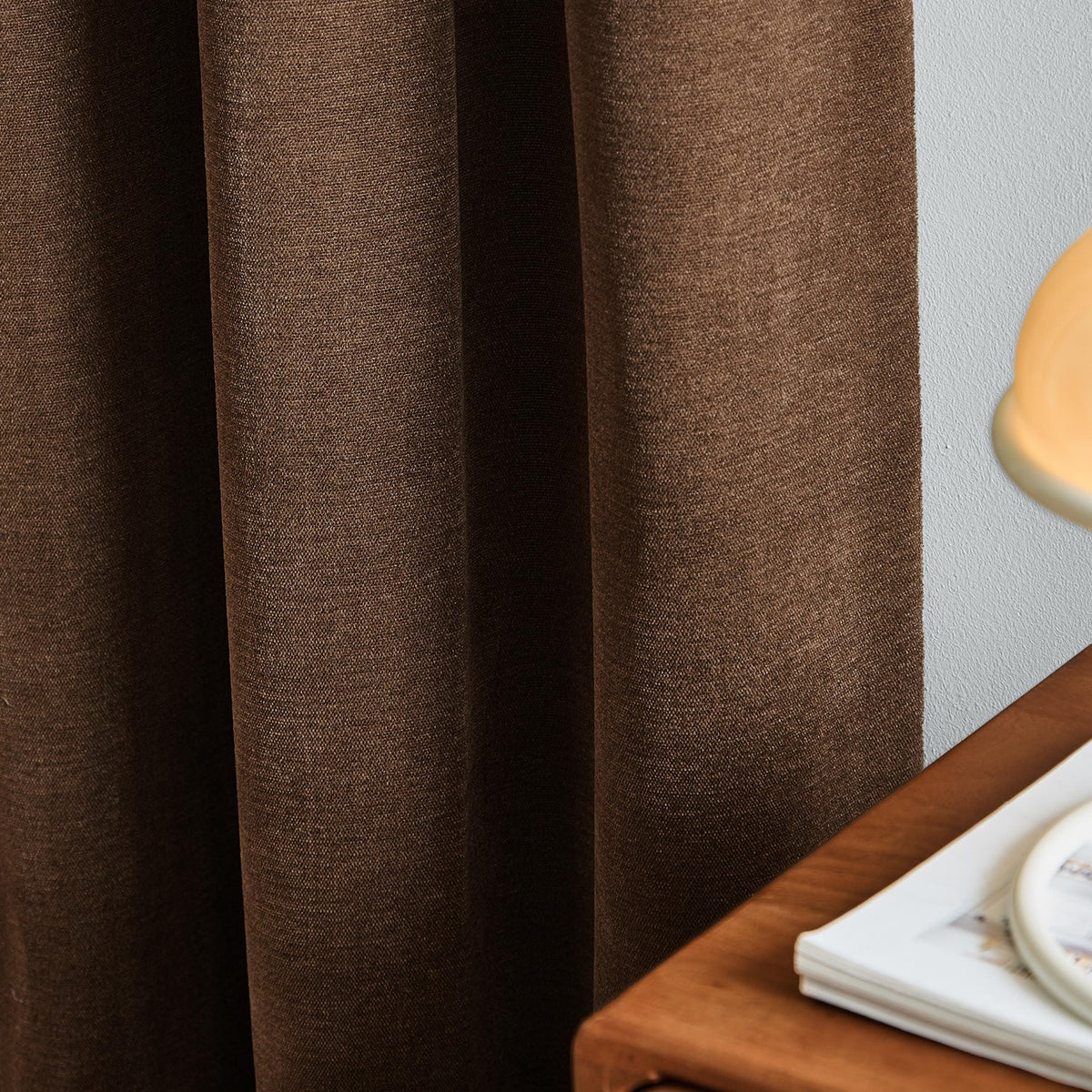 Soft chenille lined eyelet curtains in deep chocolate brown, shown draped in soft folds beside a light wall, wooden table, and lamp base.