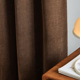 Soft chenille lined eyelet curtains in deep chocolate brown, shown draped in soft folds beside a light wall, wooden table, and lamp base.