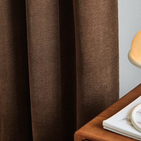 Soft chenille lined eyelet curtains in deep chocolate brown, shown draped in soft folds beside a light wall, wooden table, and lamp base.