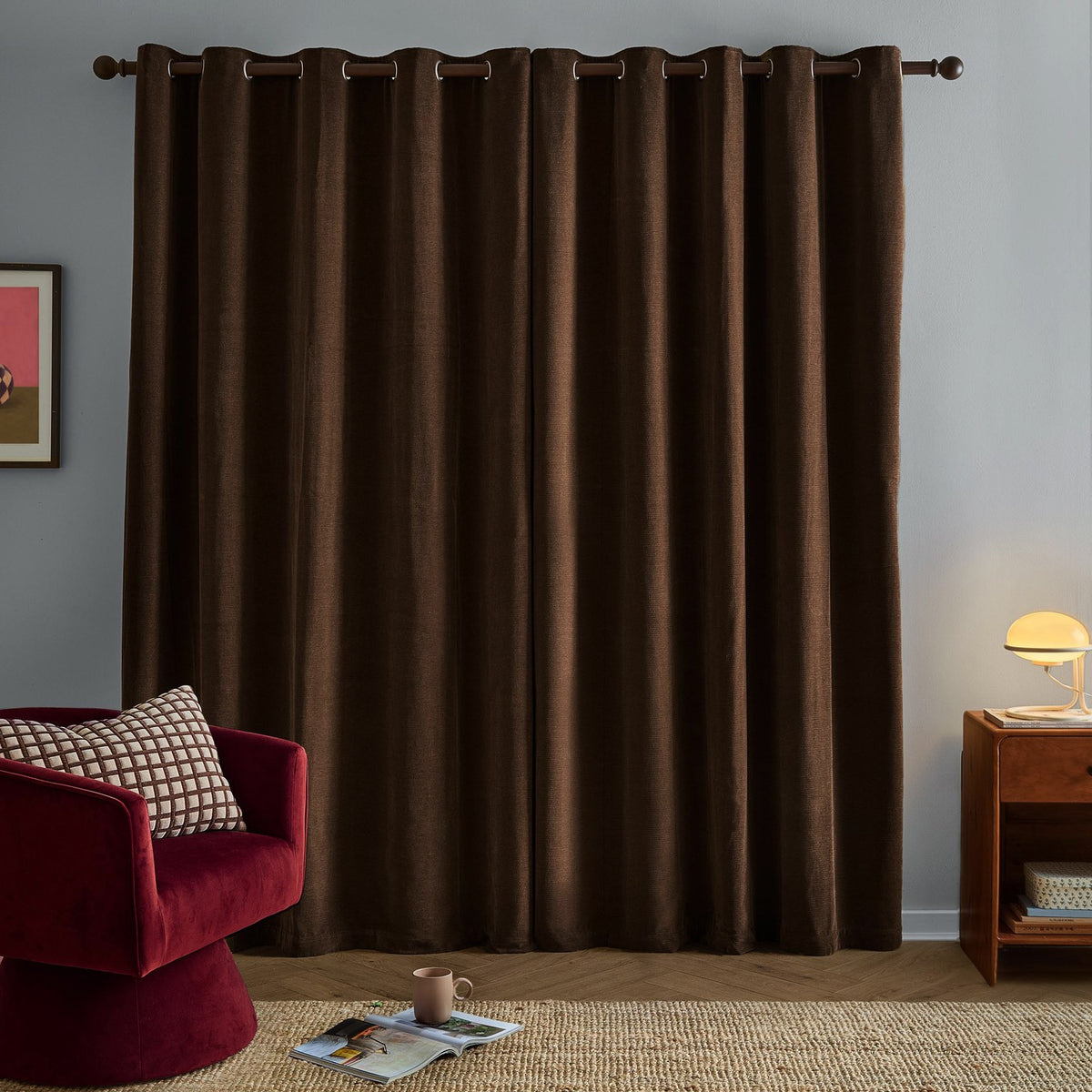 Chocolate brown chenille eyelet curtains hanging on a rod in a living room, shown with a red armchair, wooden side table, and framed wall art.