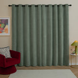 Green eyelet curtains in soft chenille fabric hung on a black rod, shown in a living room with a red armchair, patterned cushion, and framed wall art.