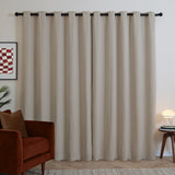 Natural oatmeal chenille eyelet curtains with dark grommets hung on a black rod, shown in a living room beside a rust-coloured armchair and side table.