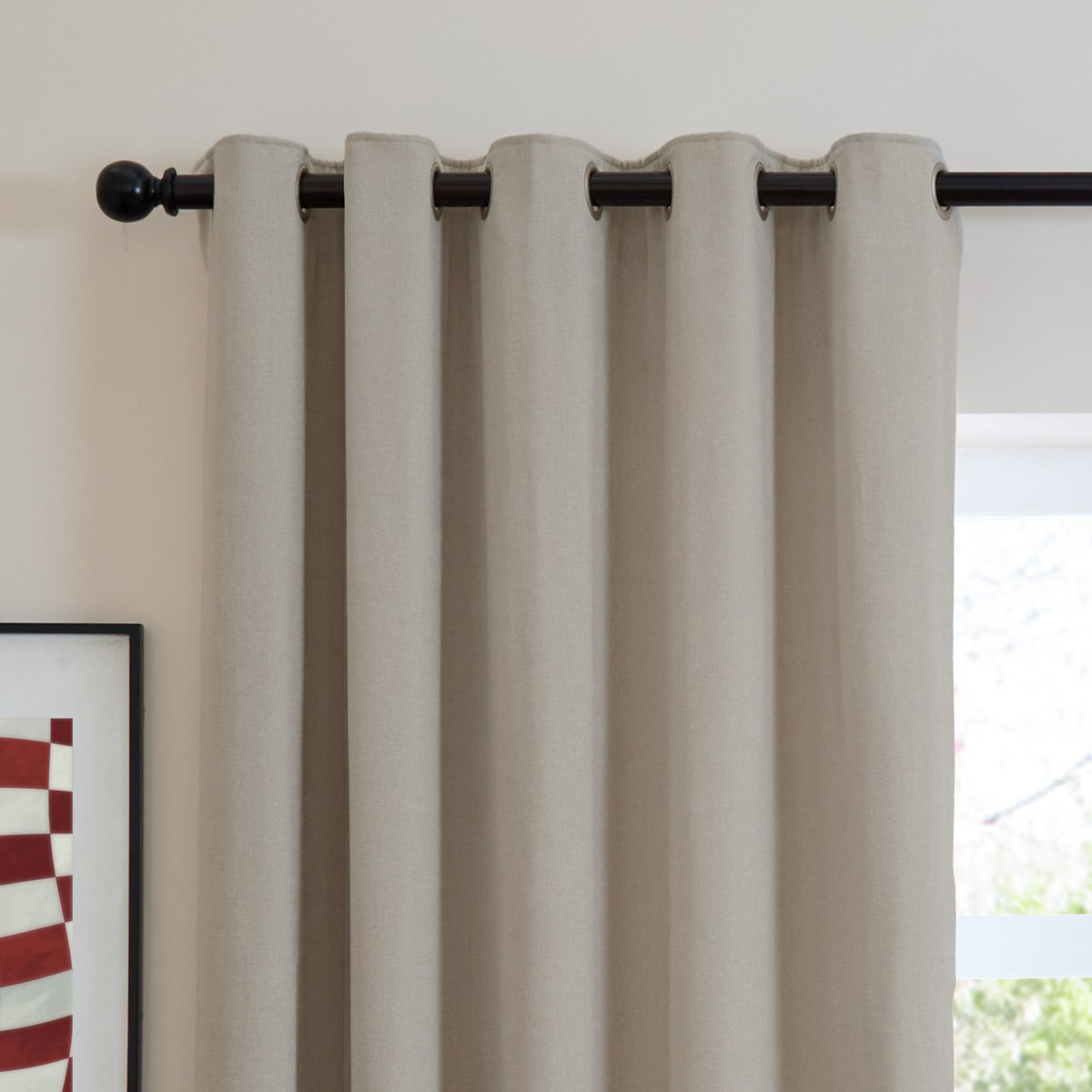 Natural oatmeal chenille eyelet curtain with metal grommets on a black rod, shown against a light wall beside a window in a living space.