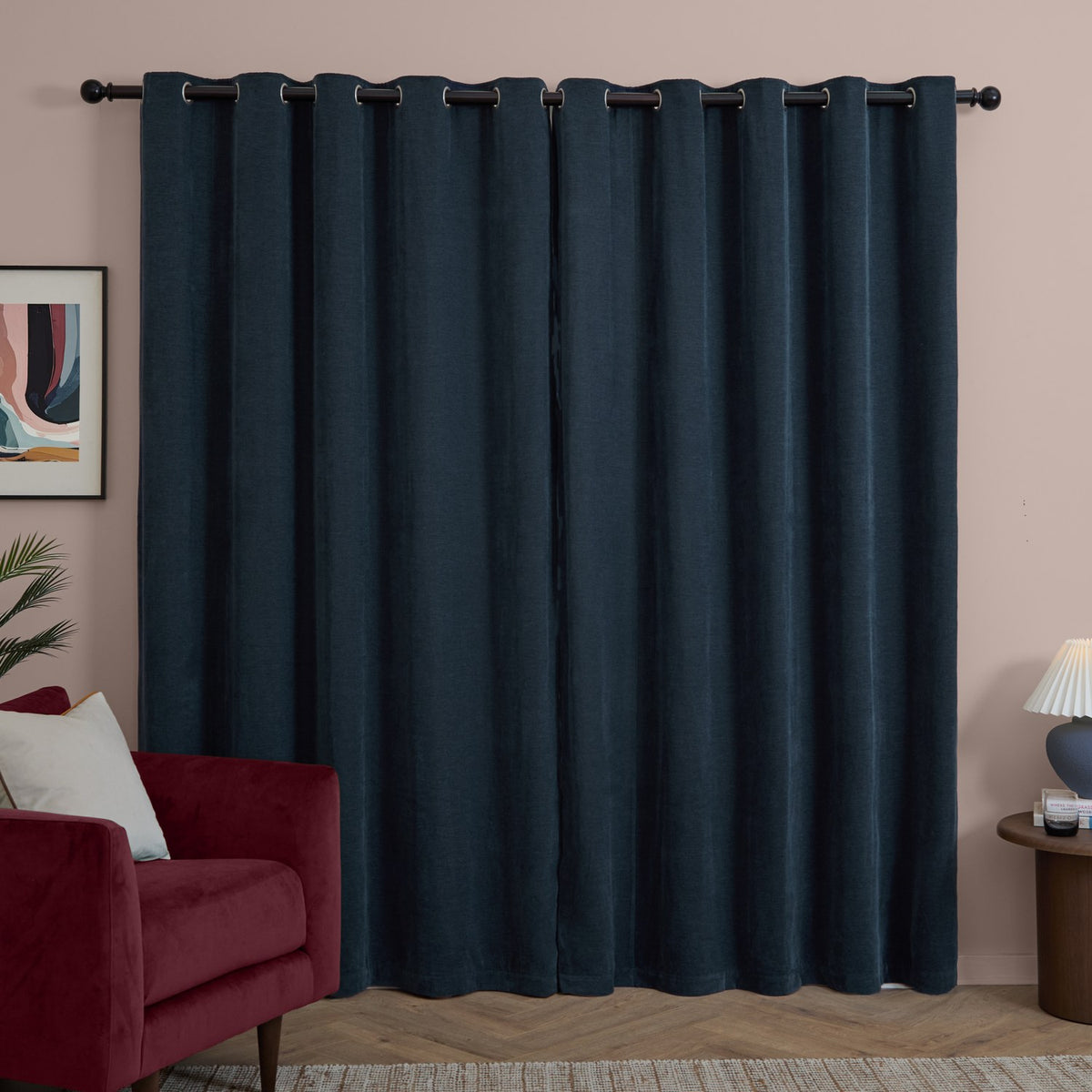 Navy chenille eyelet curtains in a pair hang from a black rod with matte grommets, shown in a living room beside a red velvet armchair and pink wall.