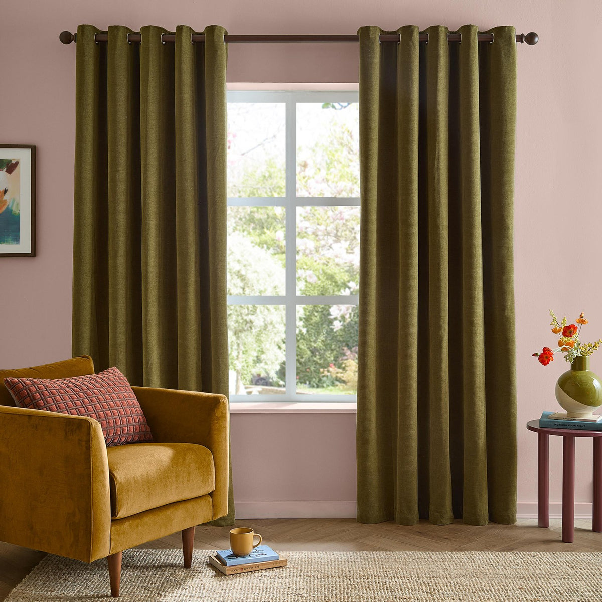 Olive green eyelet curtains in soft chenille fabric hanging at a living room window, shown with a mustard armchair and pink wall in the background.