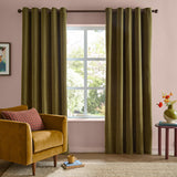 Olive green eyelet curtains in soft chenille fabric hanging at a living room window, shown with a mustard armchair and pink wall in the background.