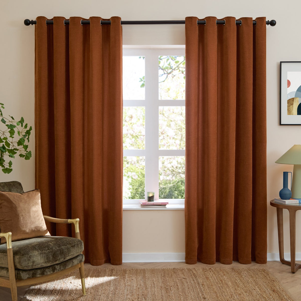 Rust chenille eyelet curtains with grommet header hung either side of a living room window, shown with an armchair, side table and framed artwork.