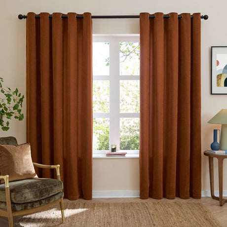 Rust chenille eyelet curtains with grommet header hung either side of a living room window, shown with an armchair, side table and framed artwork.