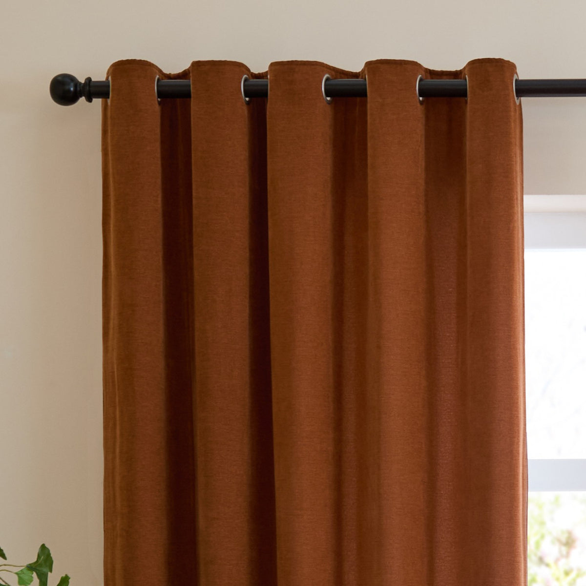 Rust chenille eyelet curtain with metal grommets on a black rod, shown in soft folds against a light wall beside a window.