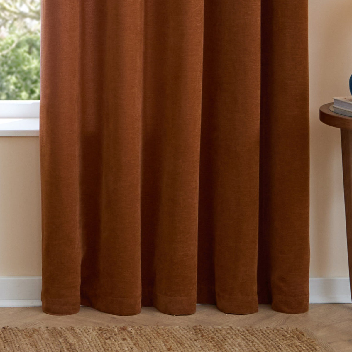 Rust chenille eyelet curtains hanging in soft folds beside a window, shown lined in a living room with a jute rug and wooden side table.