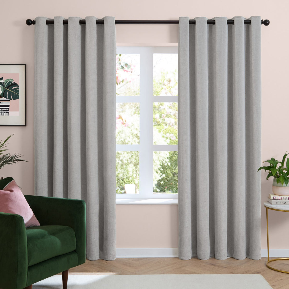 Silver grey eyelet curtains in soft chenille fabric, hung on a black pole either side of a window in a living room with a green armchair and pink walls.