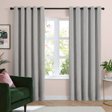 Silver grey eyelet curtains in soft chenille fabric, hung on a black pole either side of a window in a living room with a green armchair and pink walls.