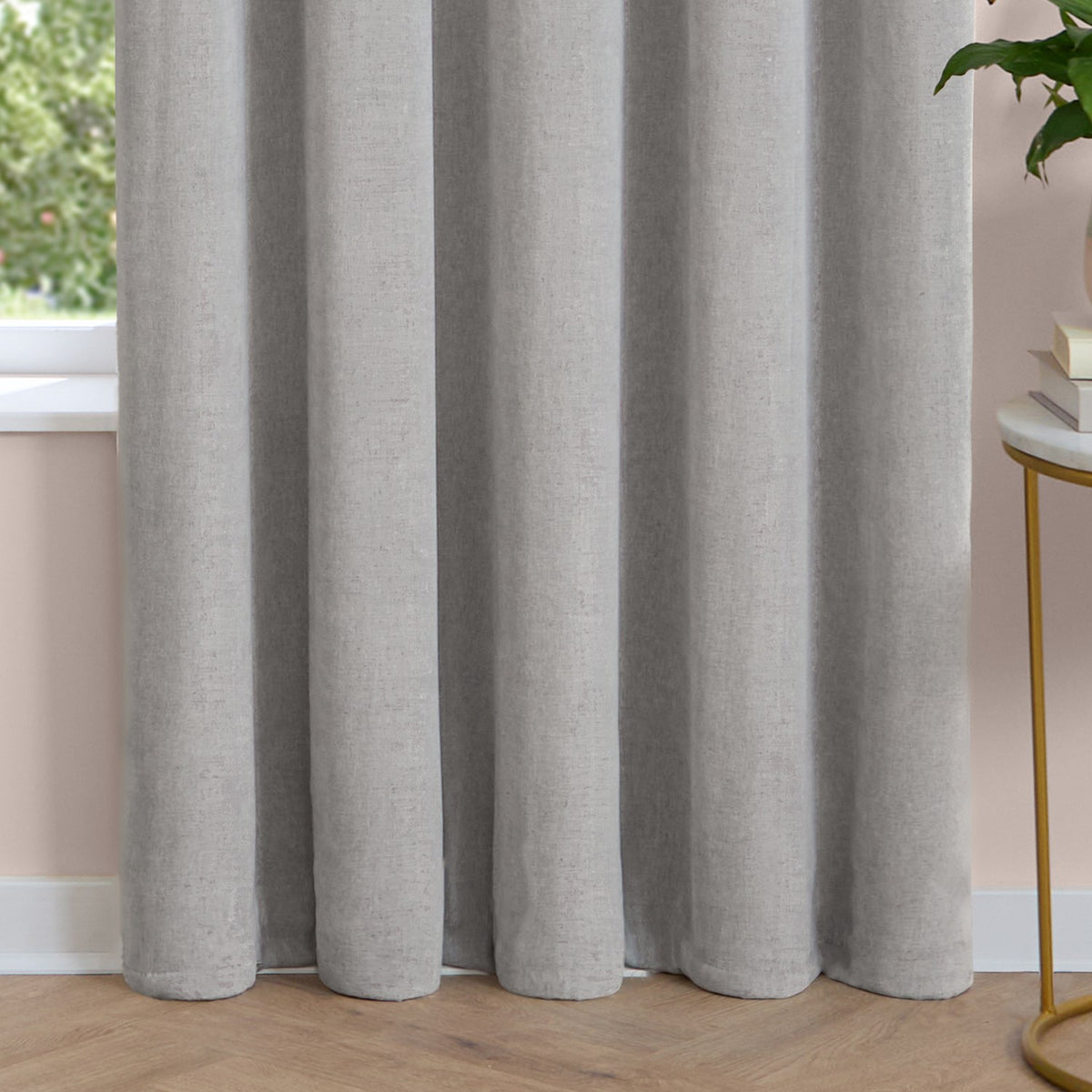 Silver grey chenille eyelet curtain with soft, lined fabric falling in deep folds, shown pooling on a wooden floor beside a window in a living room.