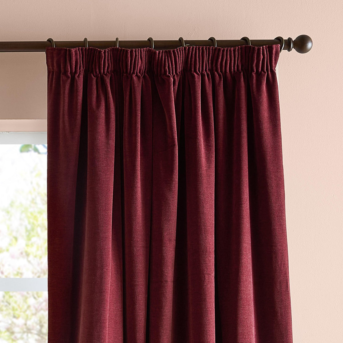 Beetroot red pencil pleat curtains in soft chenille fabric, shown hanging on a dark metal pole with ring clips against a pale pink wall near a window.