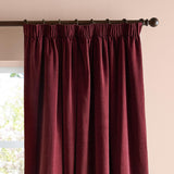 Beetroot red pencil pleat curtains in soft chenille fabric, shown hanging on a dark metal pole with ring clips against a pale pink wall near a window.