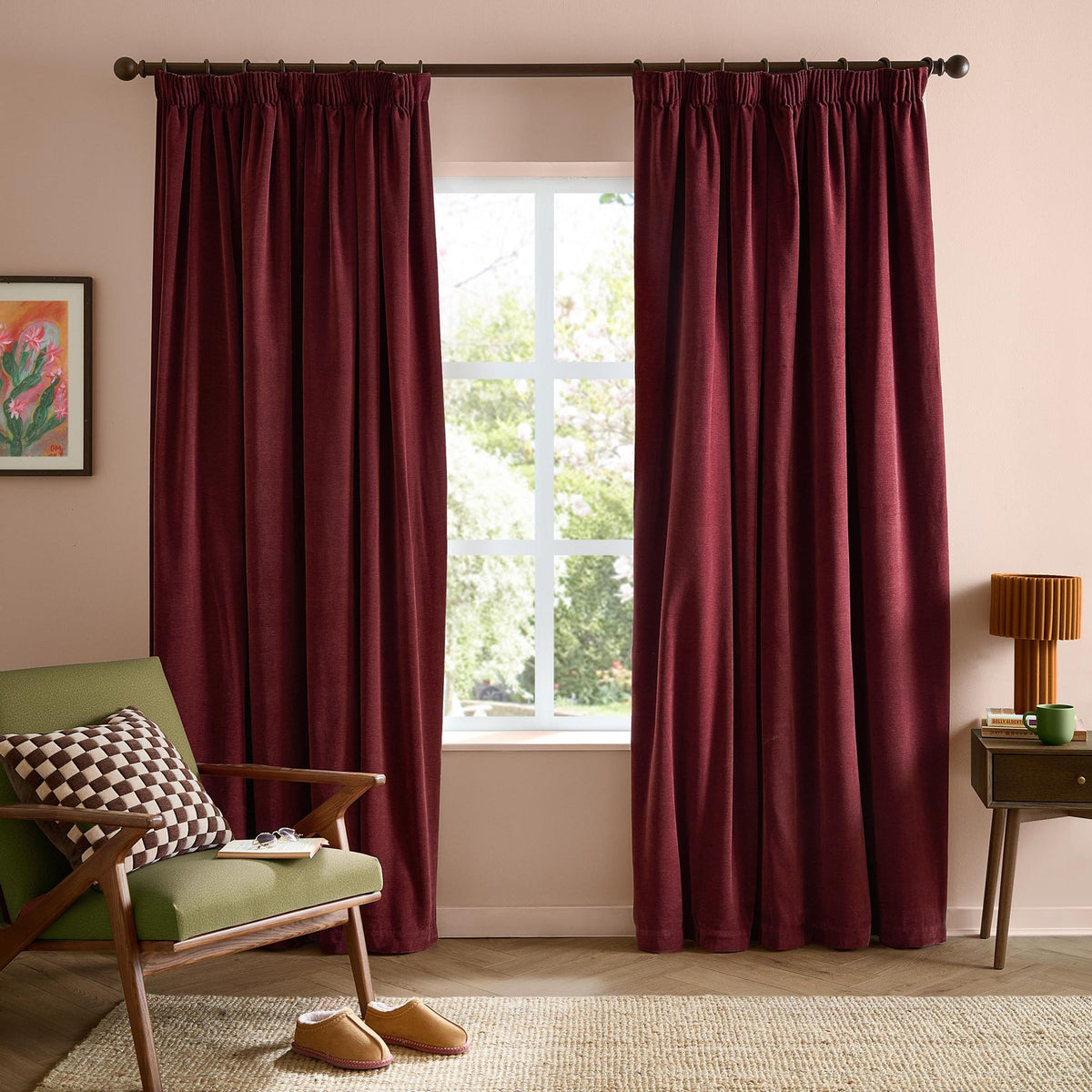 Beetroot red pencil pleat curtains in soft chenille fabric hung on a dark rod, shown in a living room with pink walls, a green armchair and window view.