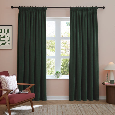 Bottle green chenille pencil pleat curtains with tape heading, shown pair-hung on a black rod either side of a window in a pink living room.