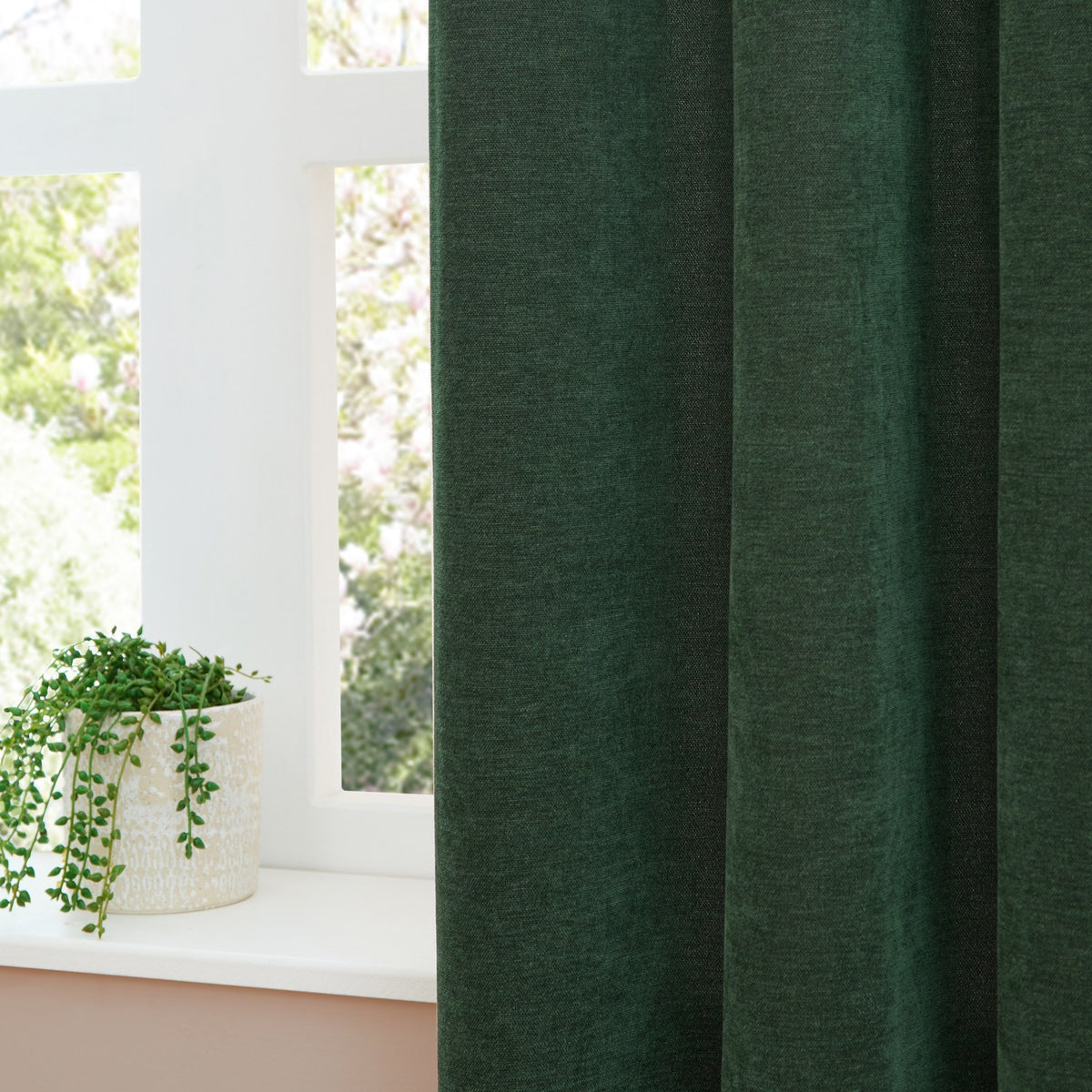 Soft Chenille Plain Polyester Pencil Pleat Curtain in bottle green, shown hanging beside a white-framed window with a potted plant on the sill.