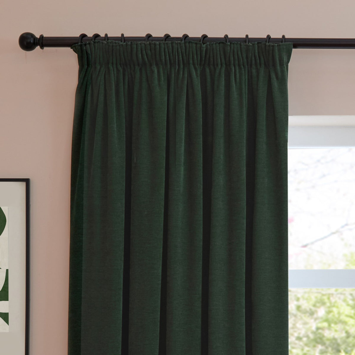 Bottle green chenille pencil pleat curtain with pencil-pleat heading on a black rod, shown hanging beside a window in a room with pink walls.