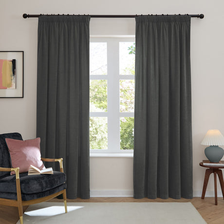 Charcoal chenille pencil pleat curtains with pencil-pleat heading hung on a black pole, shown in a living room beside an armchair and window.