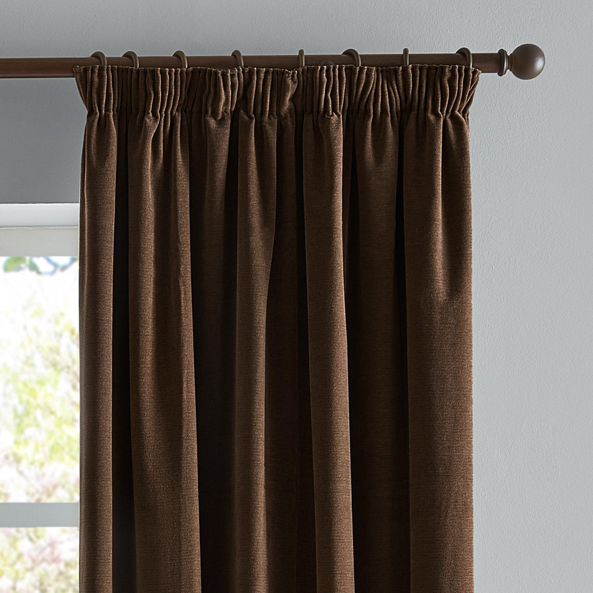 Chocolate brown chenille pencil pleat curtains with pencil-pleat heading hung on a dark metal pole, shown against a light grey wall beside a window.