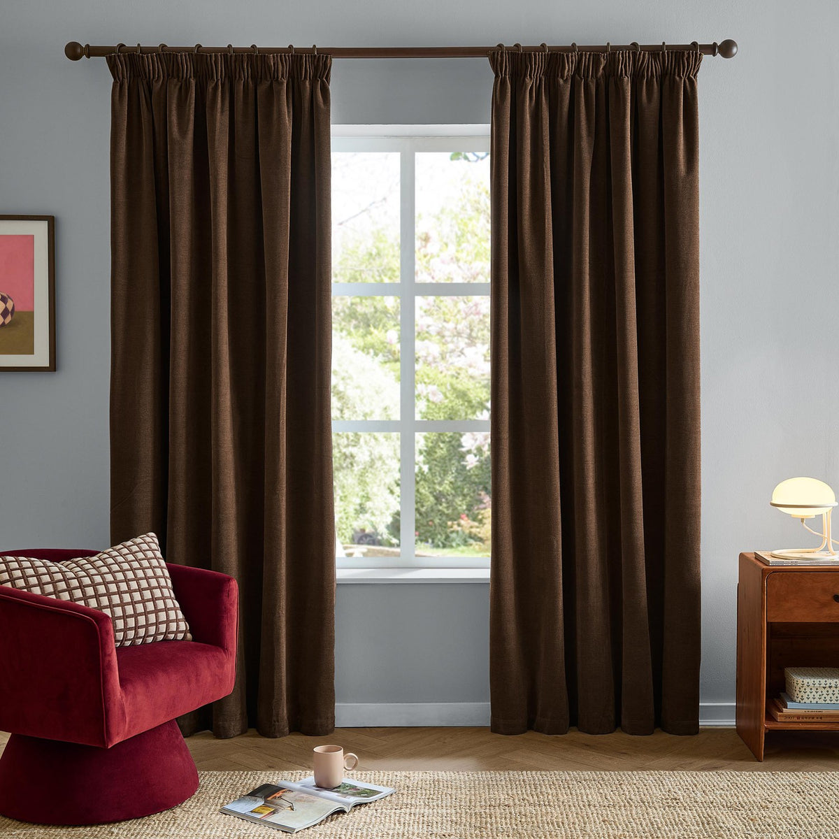 Chocolate brown chenille pencil pleat curtains hanging either side of a window in a living room, styled with a red velvet chair and wooden side table.