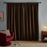 Chocolate brown chenille pencil pleat curtains hanging on a black rod in a living room, shown with a red velvet chair, wooden side table, and lamp.