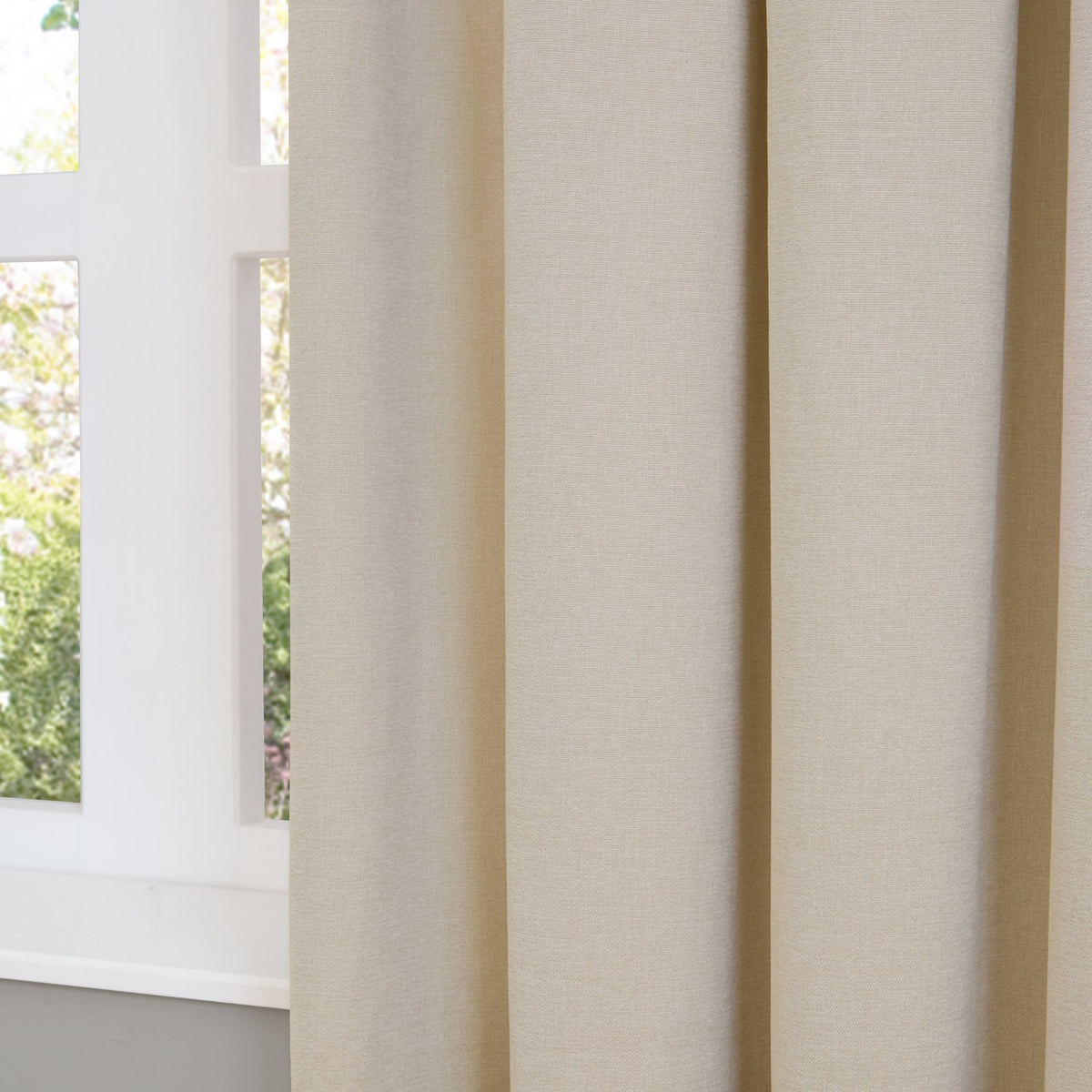 Soft chenille pencil pleat curtains in plain ecru, shown in deep folds beside a white-framed window overlooking greenery.