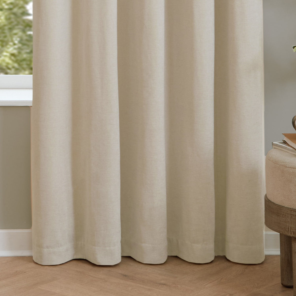 Ecru pencil pleat curtains in soft chenille fabric, shown in full-length folds beside a window in a living room with wood flooring.