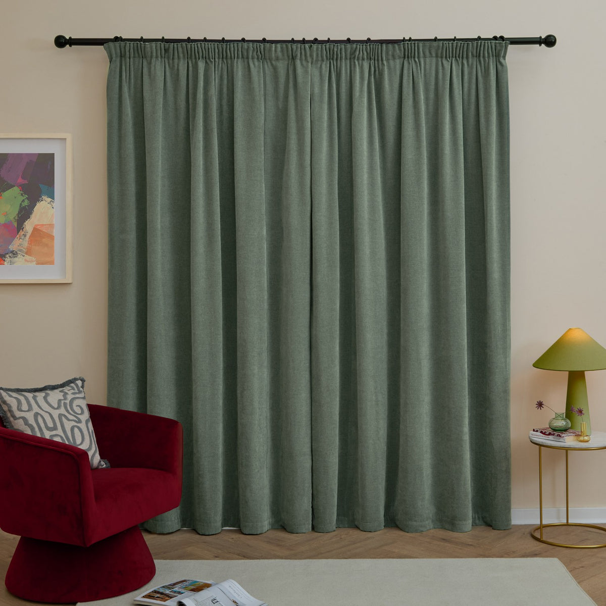 Green chenille pencil pleat curtains with pencil pleat heading on a black rod, shown floor-length in a living room beside a red armchair and side table lamp.