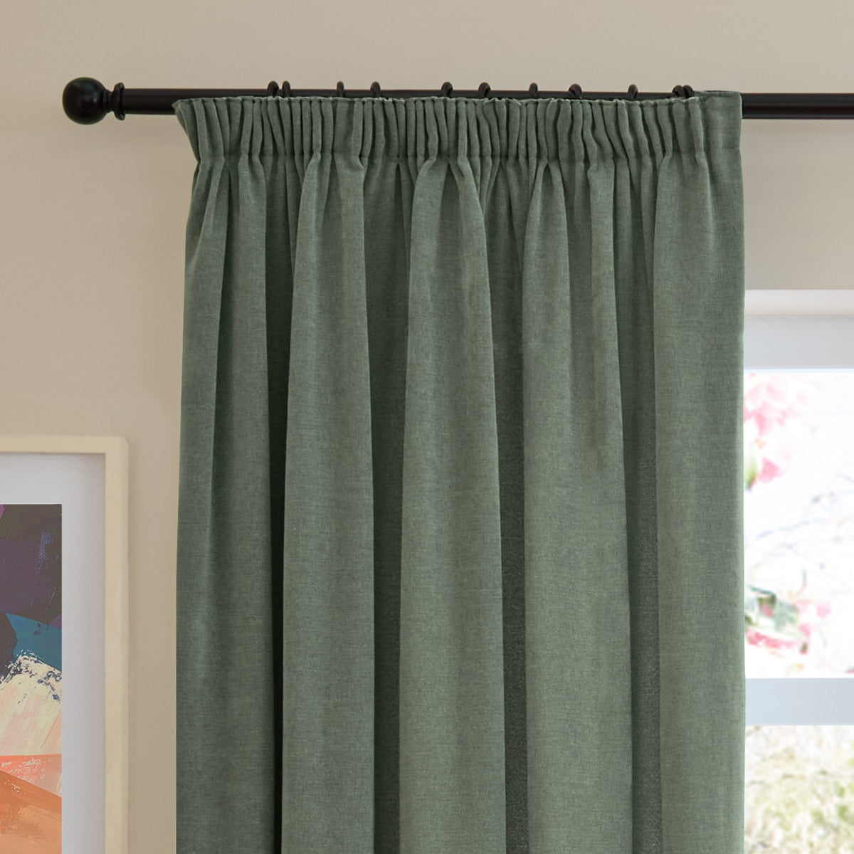 Green pencil pleat curtain in soft chenille fabric with pleated heading on a black curtain rod, shown in a room beside a window and framed artwork.