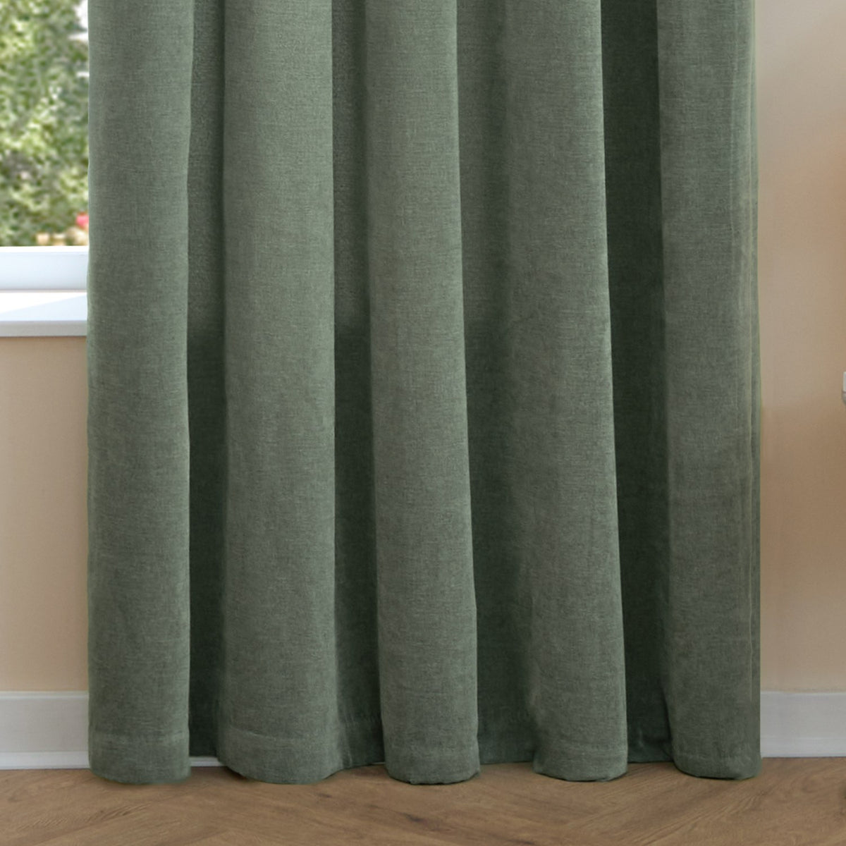 Green pencil pleat curtains in soft chenille fabric with pinch-pleated heading, shown hanging against a window in a room with wooden flooring.