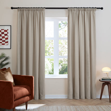 Natural beige chenille pencil pleat curtains hung on a black rod either side of a window in a living room with an armchair and side table.
