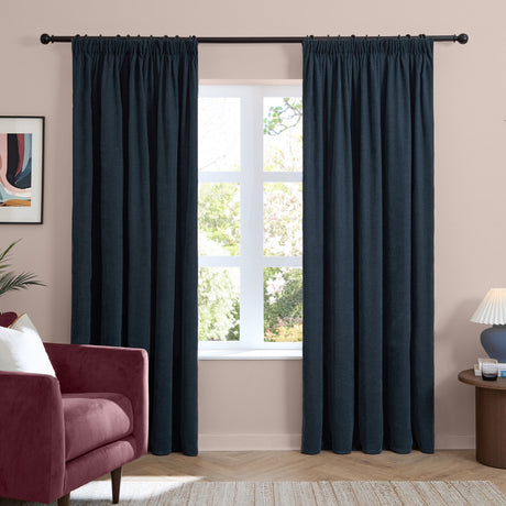 Navy chenille pencil pleat curtains hung either side of a window in a living room, shown with a pink wall and burgundy armchair.