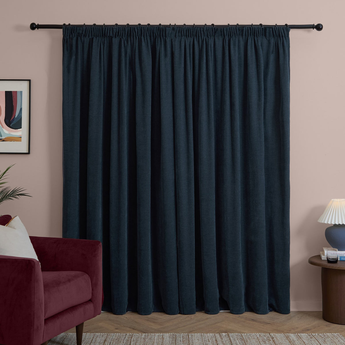 Navy chenille pencil pleat curtains with gathered heading on a black rod, shown in a living room with a burgundy velvet sofa and blush pink wall.