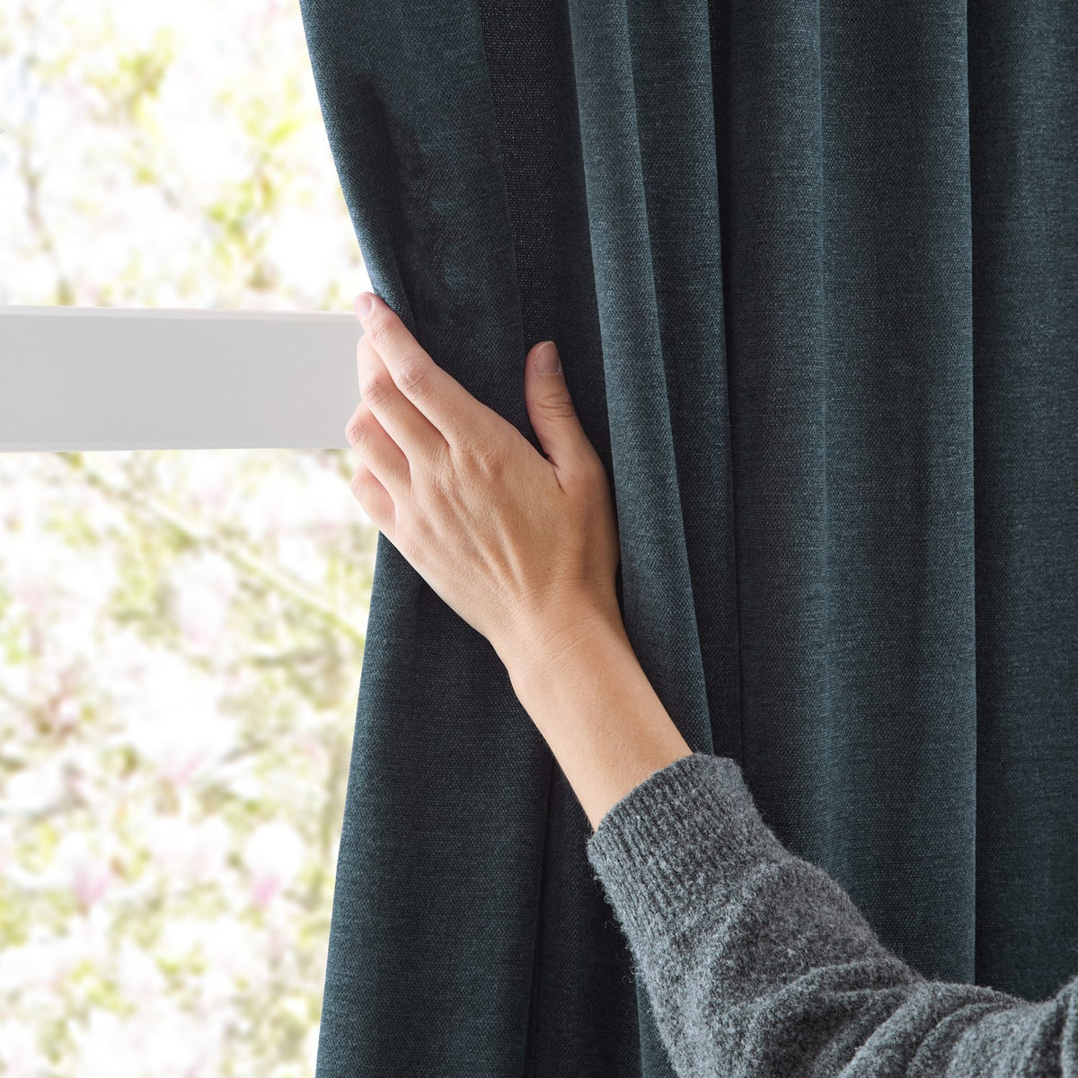 A hand adjusts a navy chenille pencil pleat curtain by a window rail, showing the fabric's nubbly pile texture against a bright, softly blurred garden view.