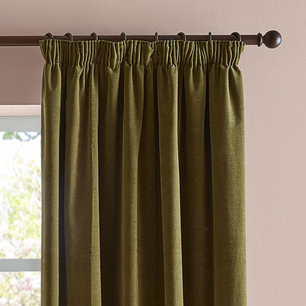 Olive green pencil pleat curtains in soft chenille fabric, shown gathered on a dark curtain rod against a pink wall near a window.