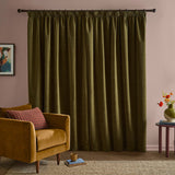 Olive green pencil pleat curtains in soft chenille fabric hanging from a rod, shown in a living room with a mustard velvet armchair and pink walls.