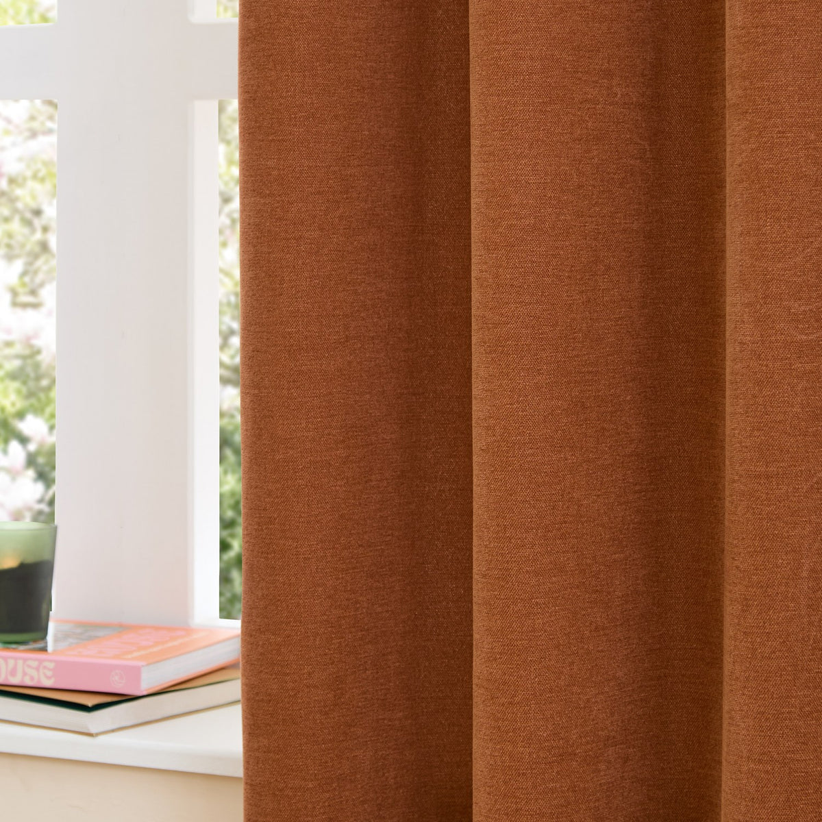 Rust chenille pencil pleat curtains in soft textured polyester, shown pleated at a white-framed window beside a stack of books in a living room setting.
