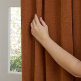 A hand pulls back a rust-coloured pencil pleat curtain in soft chenille fabric, showing the pleated heading beside a bright window.