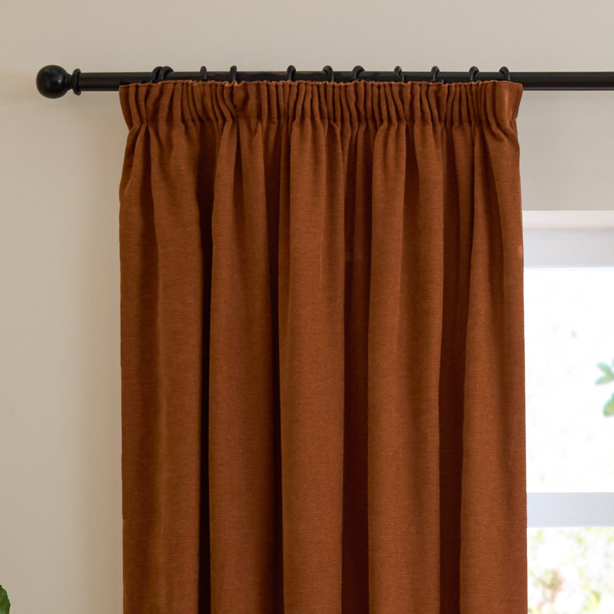 Rust orange pencil pleat curtains in soft chenille fabric, shown hanging on a black curtain pole with pencil pleat heading tape, in front of a window.