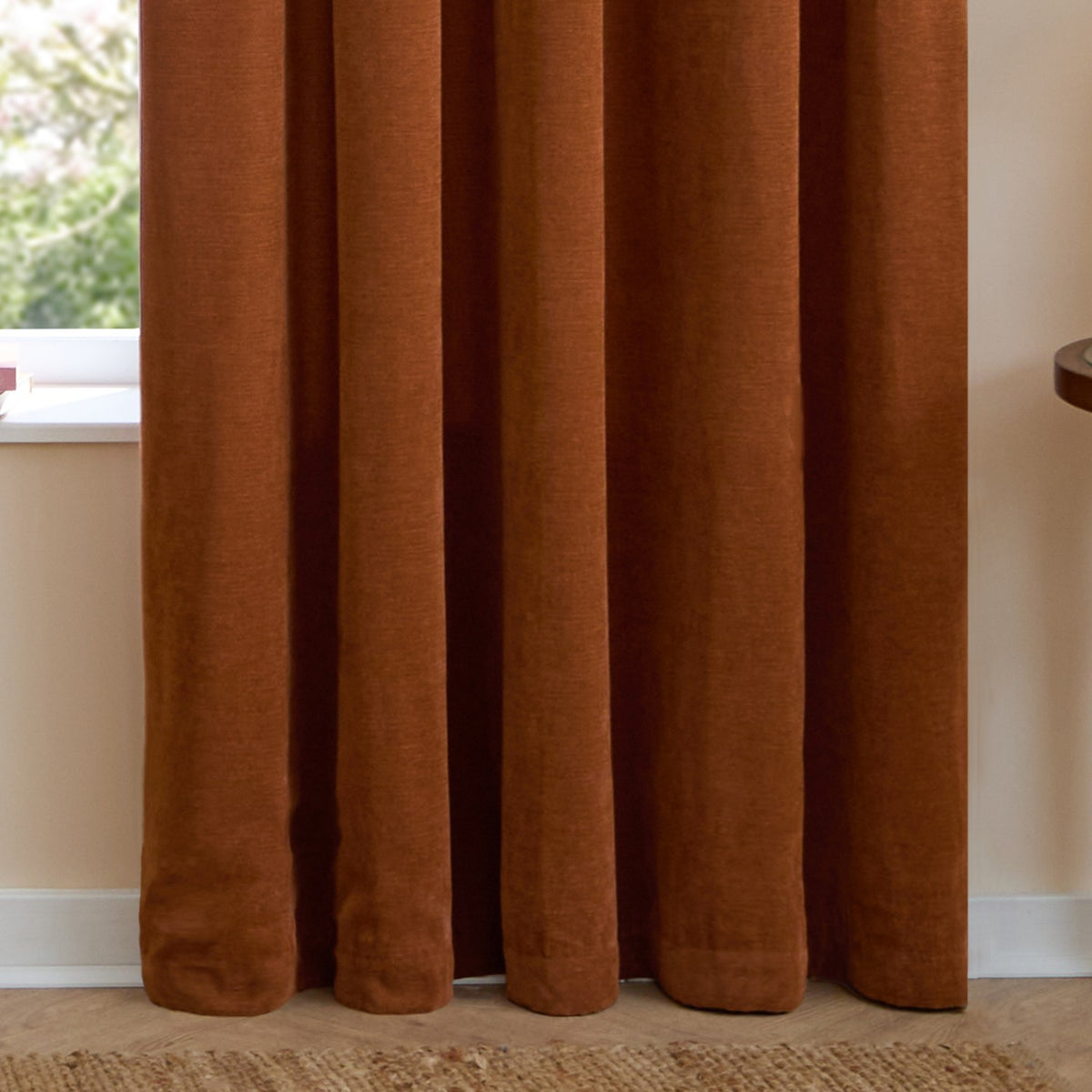 Rust chenille pencil pleat curtains with pencil-pleat heading, shown hanging in soft folds beside a window in a neutral-toned room.