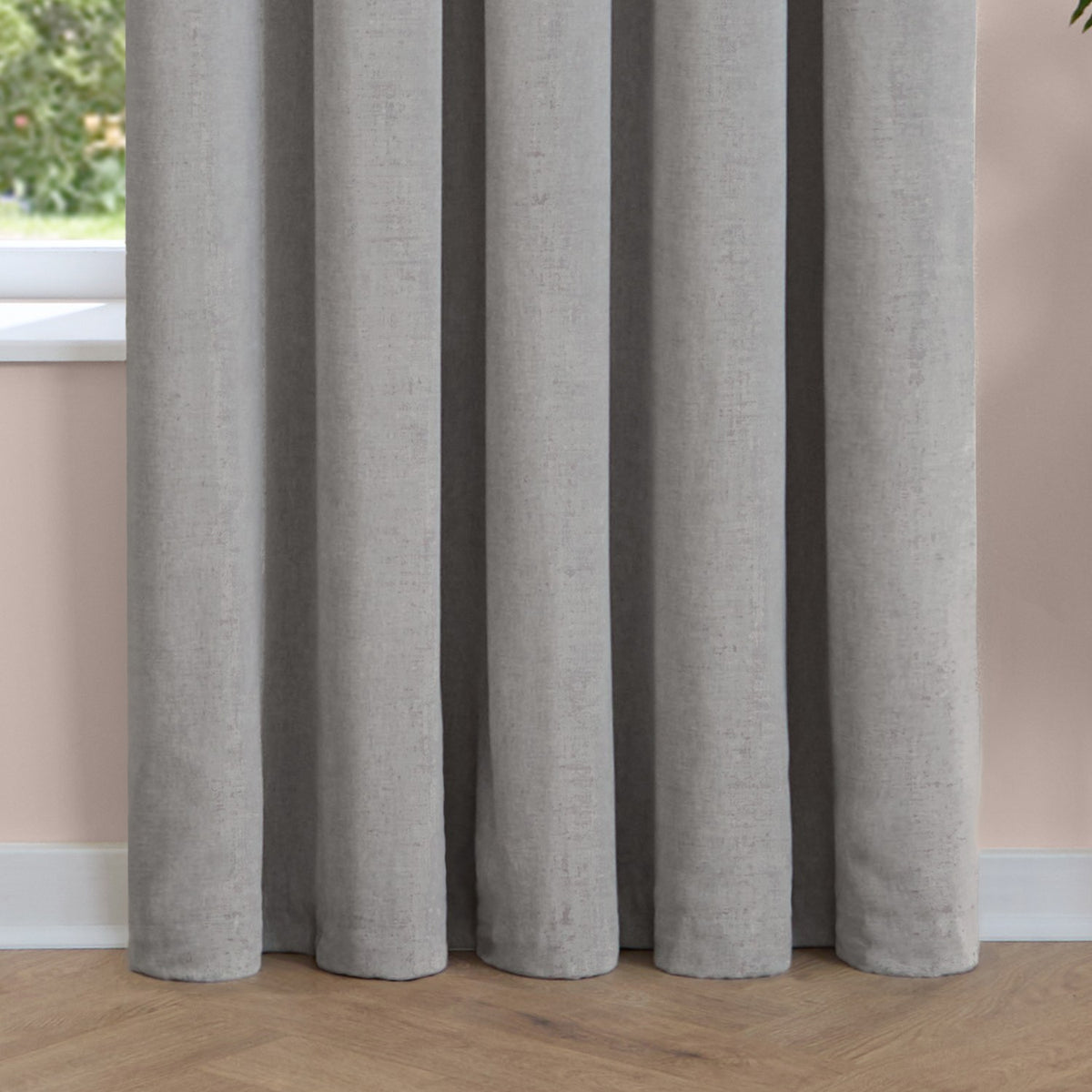 Silver-grey pencil pleat curtains in soft chenille fabric, shown pooling on a wooden floor against a pale pink wall in a room setting.