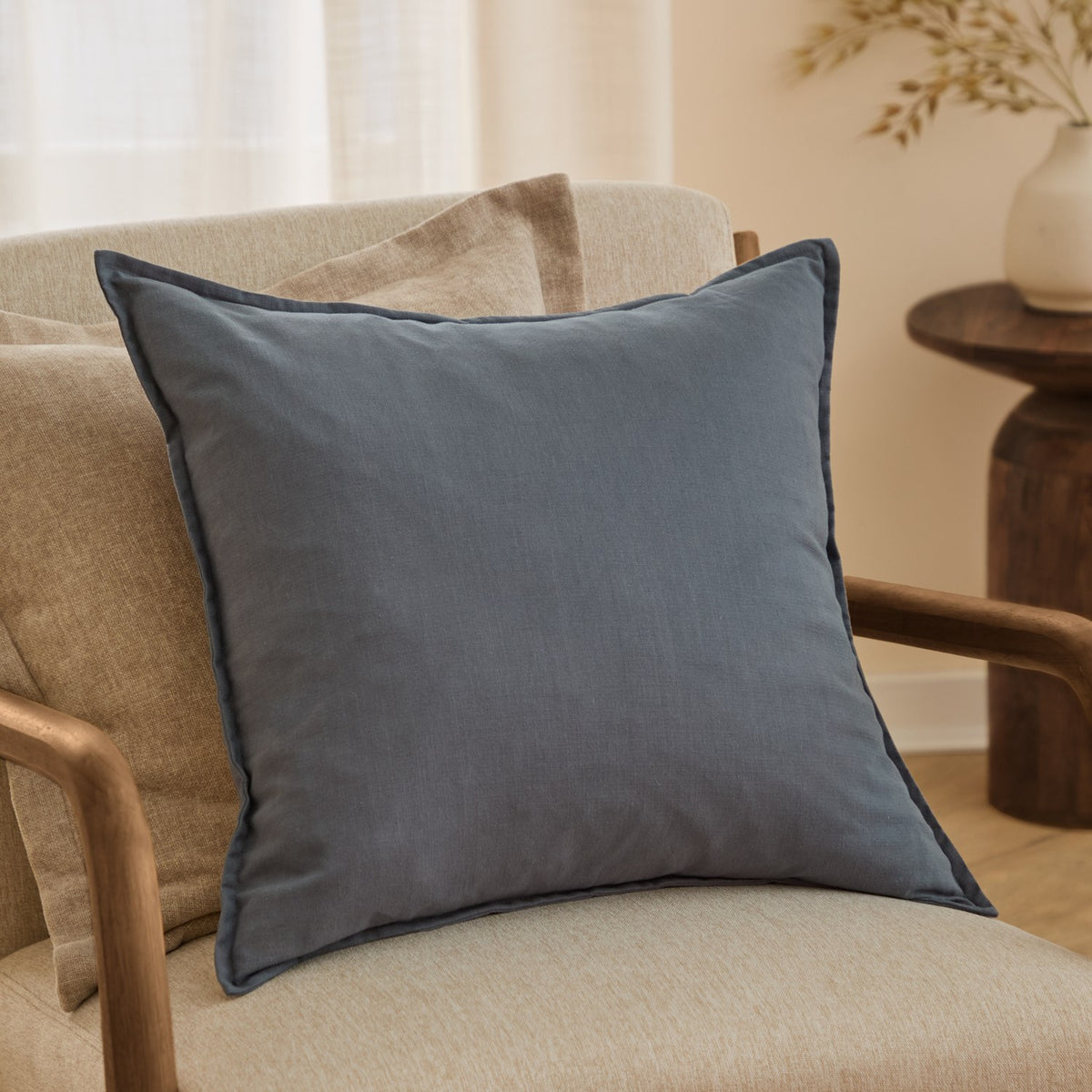 Denim blue Slub Cotton Woven Plain Cushion with flanged edge, shown on a beige armchair in a living room setting with a wooden side table.