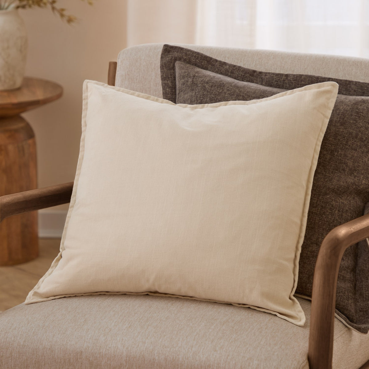 Slub Cotton Plain Linen Cushion in cream, square with flanged edges, shown on a beige armchair with a dark brown patterned cushion behind it.