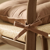 Close-up of a toffee brown slub cotton seat pad tied with fabric ties to a wooden chair leg, shown over a woven seat and cushion.
