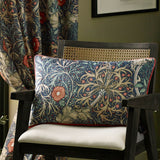 Seaweed Floral Velvet Cushion in indigo, rectangular bolster shape with printed coral floral and leaf design, shown on a dark wooden chair beside a matching patterned curtain.
