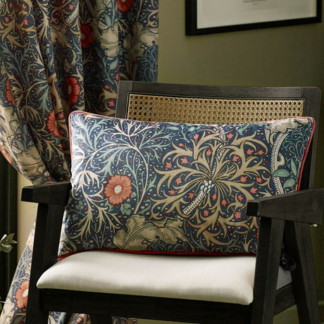Seaweed Floral Velvet Cushion in indigo, rectangular bolster shape with printed coral floral and leaf design, shown on a dark wooden chair beside a matching patterned curtain.