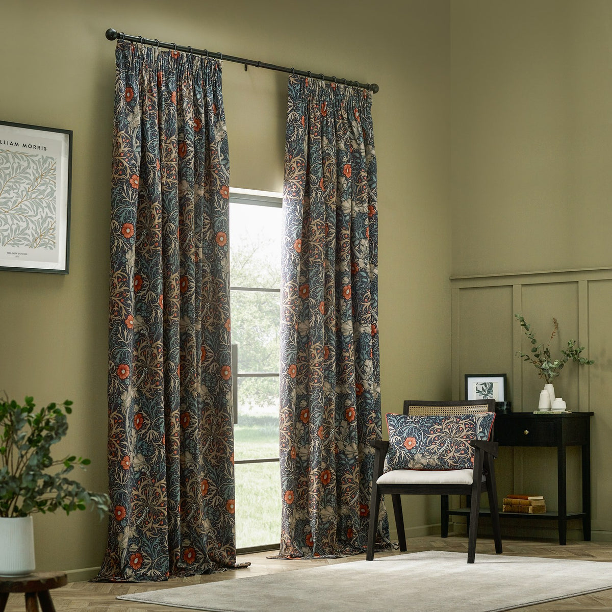 Seaweed Floral Velvet Cushion in indigo on a matching armchair, showing coral-red florals and leaf motifs, in a living room with printed curtains and dark furniture.