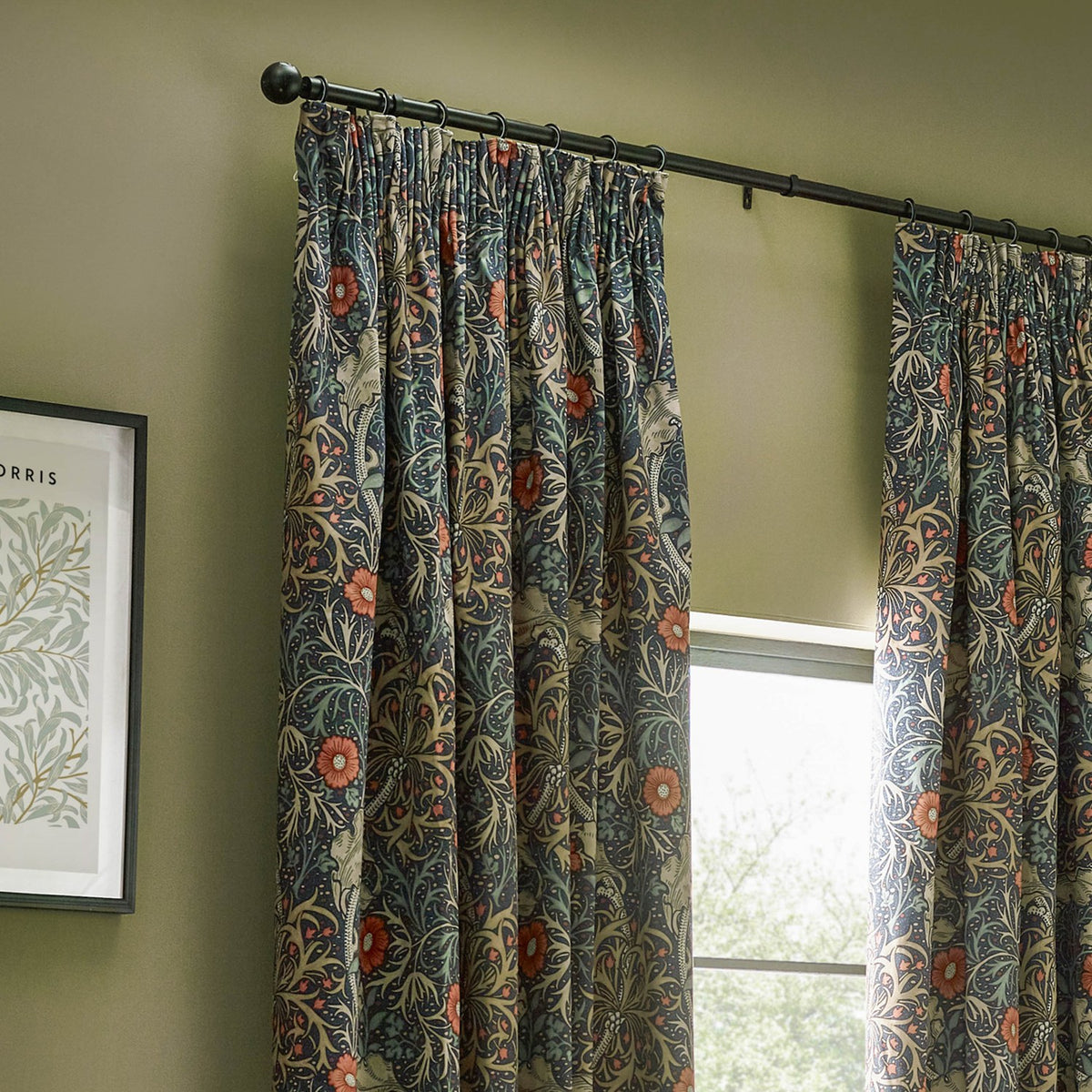 Pencil pleat curtains in indigo velvet with the Seaweed floral pattern in green, sand and coral, hung on a black rod beside a window in a green-walled room.